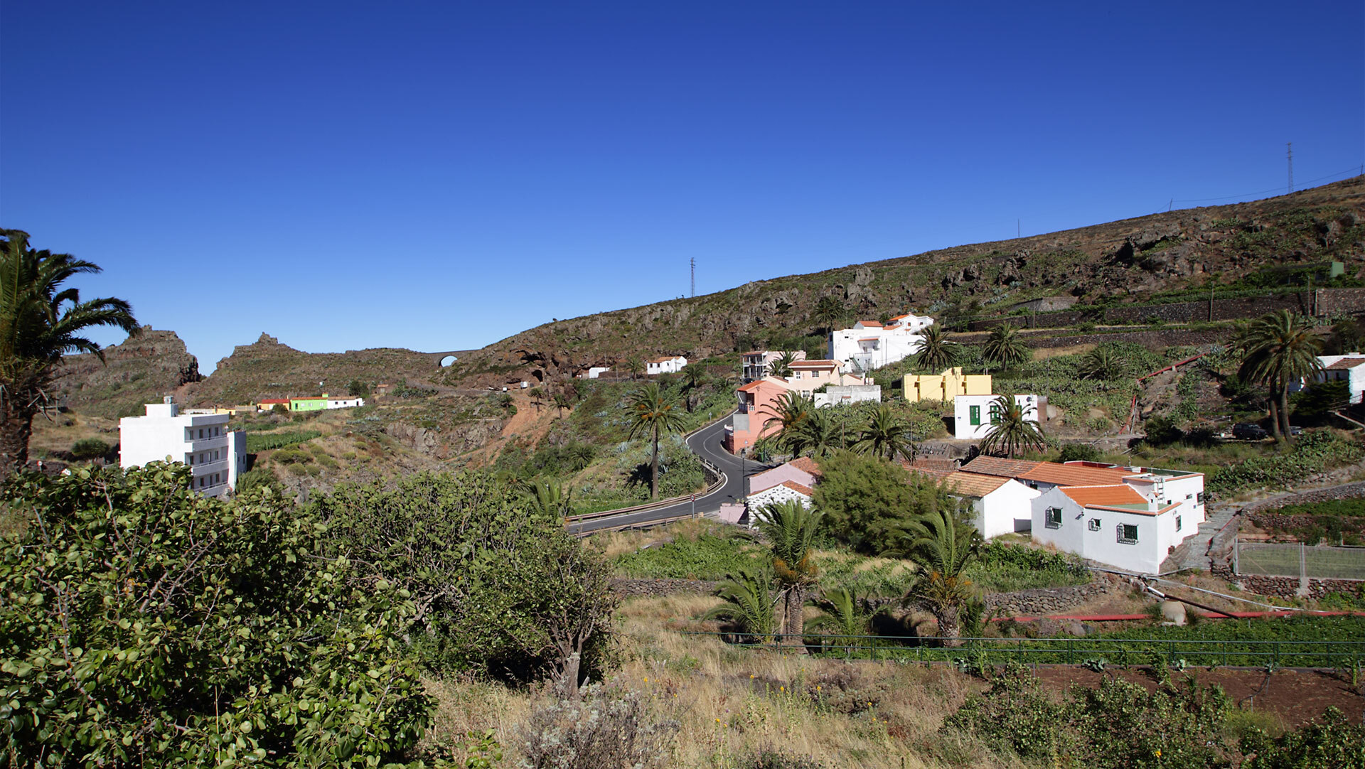 Blick auf das Hochlanddorf Arure mit seinen farbenfrohen Häusern, Palmen und der kurvigen Zufahrtsstraße durch das karge Hochland.