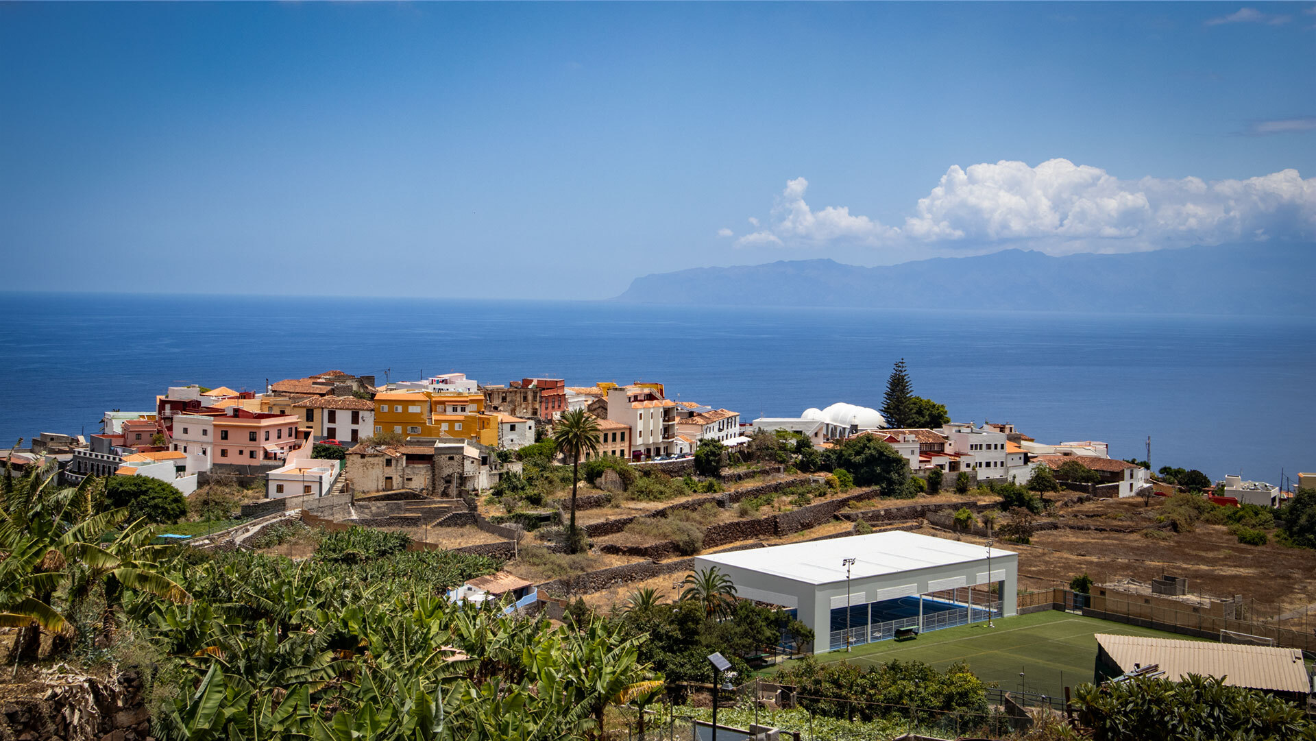 Blick auf das farbenprächtige Dorf Agulo auf La Gomera, mit Bananenplantagen im Vordergrund und der Silhouette Teneriffas am Horizont.
