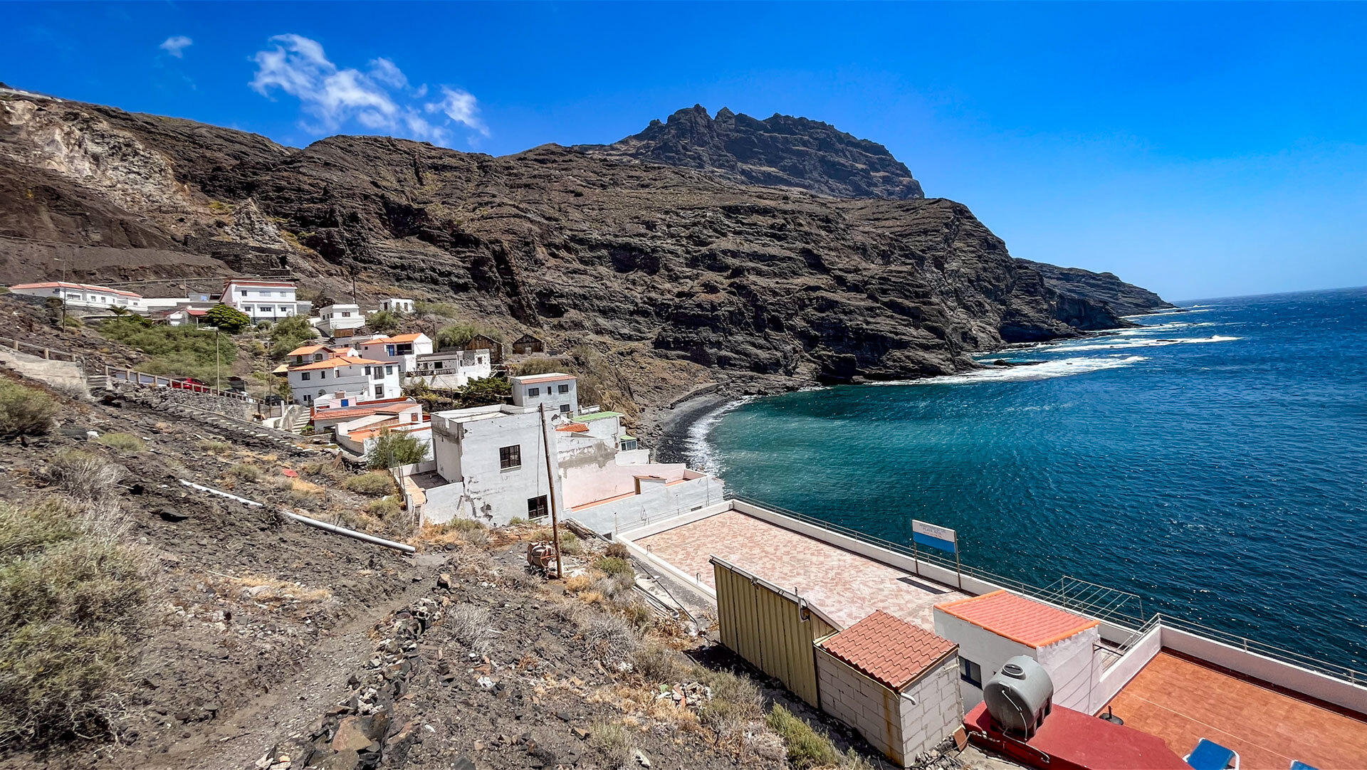 Die kleine Siedlung an der Playa de Alojera am Berghang über einer türkisblauen Bucht, überragt von schroffen Felsformationen.